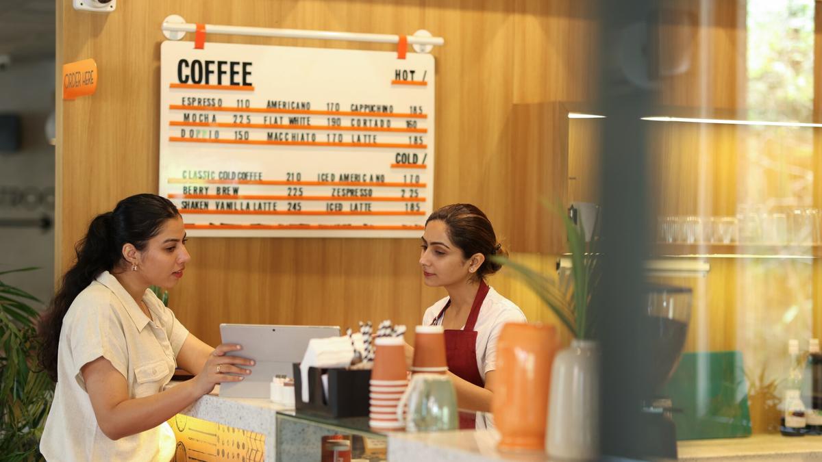 A café employee checking a shift schedule on a tablet with a customer at the counter